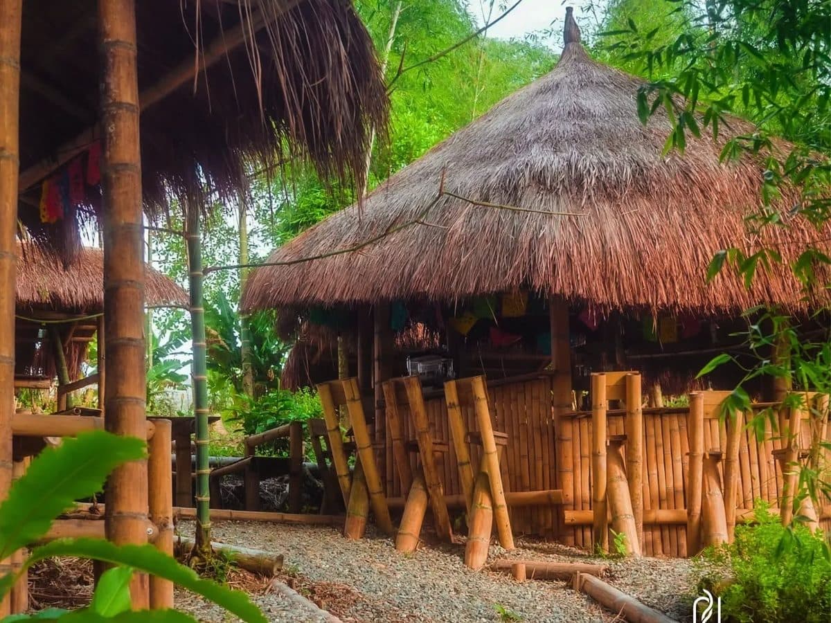 Dos quioscos de guadua con techo de paja en el jardín de Cannatel: postes y baranda de cañas de guadua, banderines de colores bajo el alero y un sendero de gravilla entre vegetación.