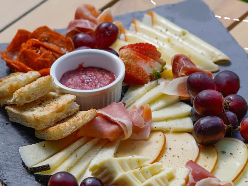 A slate board of cheeses, ham, fruit, and bread on a wooden table