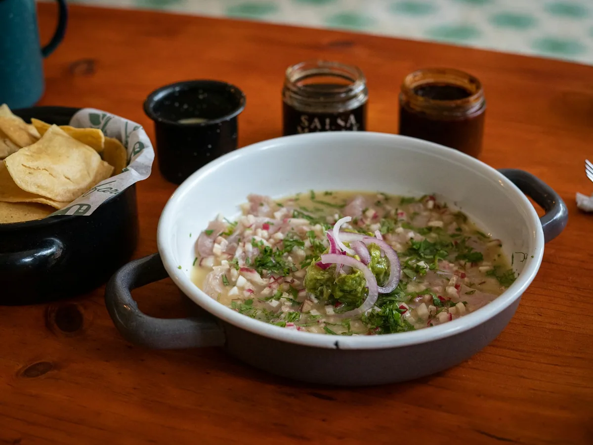 A bowl of ceviche with herbs beside totopos and sauces