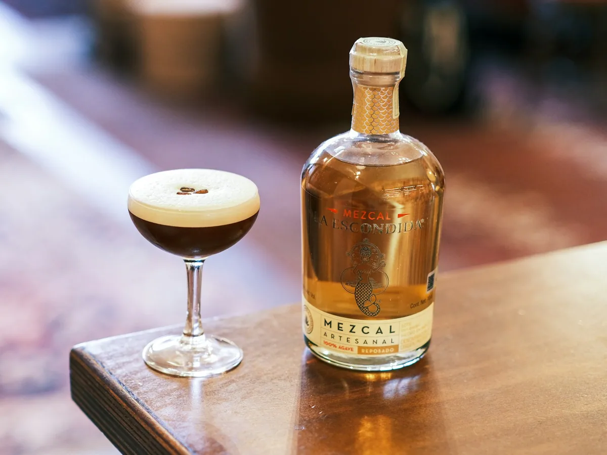 A dark cocktail in a coupe beside a bottle of mezcal
