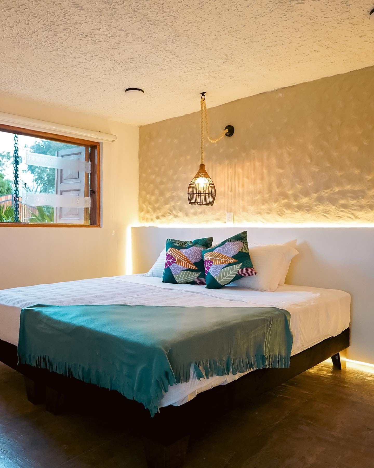 King Deluxe room with a bed dressed in green, patterned cushions, a textured wall and a window onto the garden