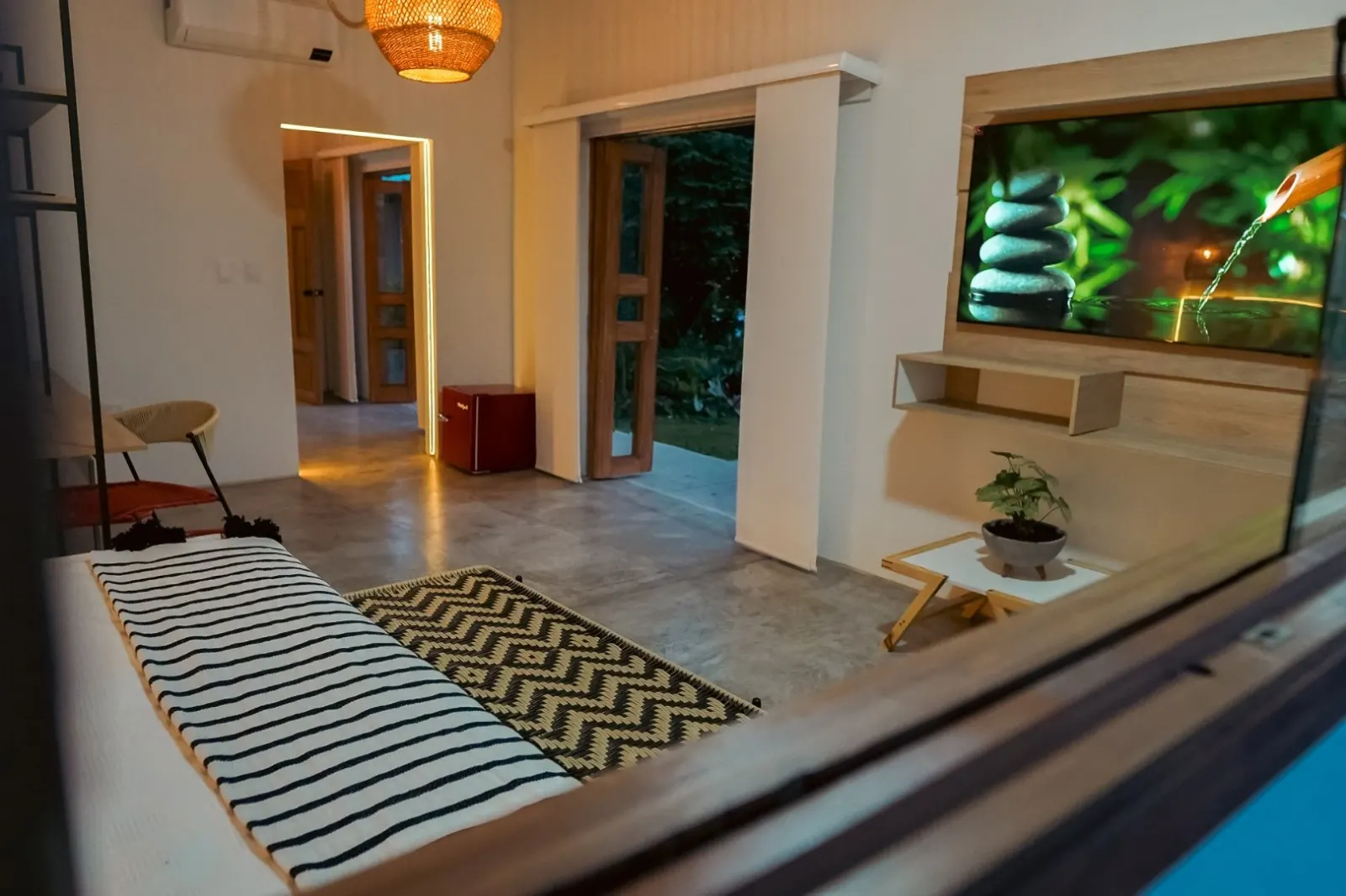 The suite’s living room seen from the window: striped daybed, zigzag rug, television, red mini fridge and wooden doors open to the garden